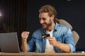 Young handsome curly smiling man with long hair in home office holding credit card and using laptop. Online shopping Royalty Free Stock Photo
