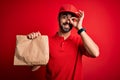 Young handsome delivery man with beard wearing cap holding takeaway paper bag with food with happy face smiling doing ok sign with Royalty Free Stock Photo