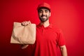 Young handsome delivery man with beard wearing cap holding takeaway paper bag with food with a happy face standing and smiling Royalty Free Stock Photo