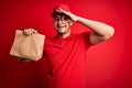 Young handsome delivery man holding paper bag with takeaway food over red background with happy face smiling doing ok sign with Royalty Free Stock Photo