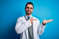 Young handsome doctor man with beard wearing coat and glasses over blue background amazed and smiling to the camera while Royalty Free Stock Photo