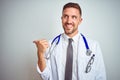 Young handsome doctor man wearing white professional coat over isolated background smiling with happy face looking and pointing to Royalty Free Stock Photo