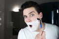 Young handsome funny man with foam on his face gmirqacing while shaving with razor in bathroom in the morning everyday routine Royalty Free Stock Photo