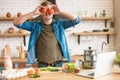 Young handsome funny man holding tomatoes near his eyes in well-equipped modern kitchen while cooking tasty breakfast , sunday