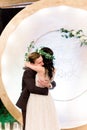 Young handsome groom in dark suit and bride in white dress and floral wreath standing near arch and kissing. Happy Royalty Free Stock Photo