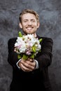 Young handsome groom smiling, holding bridal bouquet over grey background.