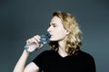Young handsome guy in a black T-shirt with long blonde hair drinks clear water from a bottle on a gray background