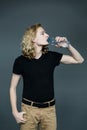 Young handsome guy in a black T-shirt with long blonde hair drinks clear water from a glass on a gray background