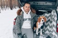 Young handsome guy in a snowy winter forest sits in the trunk of his car in an embrace with his friend Beagle breed dog Royalty Free Stock Photo