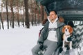 Young handsome guy in a snowy winter forest sits in the trunk of his car in an embrace with his friend Beagle breed dog Royalty Free Stock Photo