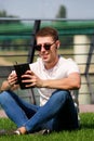 Young handsome guy with sunglasses using tablet and headphones, drinking coffee to go, man sitting on grass and enjoying sunny day Royalty Free Stock Photo