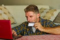 Young handsome and happy man holding credit card sitting at home sofa couch using laptop computer online shopping smiling cheerful Royalty Free Stock Photo