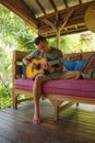 Young handsome and happy mixed ethnicity man in hipster style chilling outdoors playing guitar relaxed at tranquil tropical garden Royalty Free Stock Photo