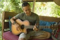 Young handsome and happy mixed ethnicity man in hipster style chilling outdoors playing guitar relaxed at tranquil tropical garden Royalty Free Stock Photo