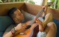 Young handsome and happy mixed ethnicity man in hipster style chilling outdoors playing guitar relaxed at tranquil tropical garden Royalty Free Stock Photo