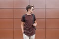 Young handsome hipster man in dark stylish sunglasses in fashionable summer clothes with a backpack poses near the brown wall Royalty Free Stock Photo