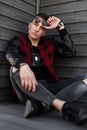 Young handsome hipster man in dark sunglasses in a trendy red checkered jacket in sneakers in gray ripped jeans is sitting