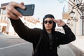 Young handsome hipster man in fashionable black clothes in a cap in sunglasses is posing and makes a selfie on a mobile phone Royalty Free Stock Photo