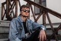 Young handsome hipster man in a fashionable denim jacket in black pants in stylish dark sunglasses relax sitting Royalty Free Stock Photo