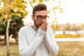 Young handsome hipster man in glasses and a white sweater is walking in a cold autumn park. Heats his hands with his breath.