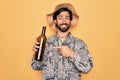 Young handsome hispanic bohemian hipster man with bear drinking from a empty wine bottle very happy pointing with hand and finger