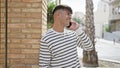 Young, handsome hispanic man confidently smiling, enjoying sunny day on city street, casually leaning against wall, talking on Royalty Free Stock Photo