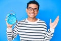 Young handsome hispanic man holding alarm clock celebrating victory with happy smile and winner expression with raised hands Royalty Free Stock Photo
