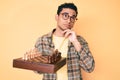 Young handsome hispanic man holding chess board wearing glasses serious face thinking about question with hand on chin, thoughtful Royalty Free Stock Photo