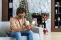 Young handsome hispanic man at home sitting on sofa in living room for Christmas near tree, man using phone happily Royalty Free Stock Photo