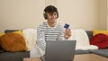 Young, handsome hispanic man smiles, listening to music with headphones and holding a credit card, sitting on living room sofa Royalty Free Stock Photo