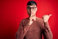 Young handsome hispanic man wearing nerd glasses over red background asking to be quiet with finger on lips pointing with hand to Royalty Free Stock Photo