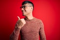 Young handsome hispanic man wearing nerd glasses over red background smiling with happy face looking and pointing to the side with Royalty Free Stock Photo