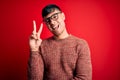Young handsome hispanic man wearing nerd glasses over red background smiling with happy face winking at the camera doing victory Royalty Free Stock Photo