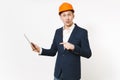 Young handsome impressed businessman in dark suit, protective hardhat pointing index finger on tablet pc computer