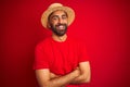 Young handsome indian man wearing t-shirt and hat over isolated red background happy face smiling with crossed arms looking at the Royalty Free Stock Photo