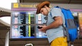 Young handsome international airport tourist passenger in hat uses mobile phone stands with backpack and suitcase at