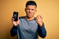 Young handsome latin man holding broken smartphone showing craked screen annoyed and frustrated shouting with anger, crazy and Royalty Free Stock Photo