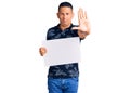 Young handsome latin man holding cardboard banner with blank space with open hand doing stop sign with serious and confident Royalty Free Stock Photo