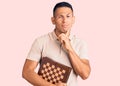 Young handsome latin man holding chess serious face thinking about question with hand on chin, thoughtful about confusing idea Royalty Free Stock Photo