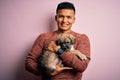 Young handsome latin man holding cute puppy pet over isolated pink background with a happy face standing and smiling with a Royalty Free Stock Photo