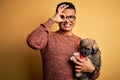 Young handsome latin man holding cute puppy pet over isolated yellow background with happy face smiling doing ok sign with hand on Royalty Free Stock Photo