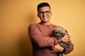 Young handsome latin man holding cute puppy pet over isolated yellow background with a happy face standing and smiling with a Royalty Free Stock Photo