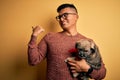 Young handsome latin man holding cute puppy pet over isolated yellow background pointing and showing with thumb up to the side Royalty Free Stock Photo