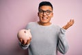 Young handsome latin man holding piggy bank over isolated pink background very happy and excited, winner expression celebrating Royalty Free Stock Photo