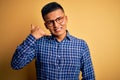 Young handsome latin man wearing casual shirt and glasses over yellow background smiling doing phone gesture with hand and fingers Royalty Free Stock Photo