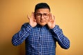 Young handsome latin man wearing casual shirt and glasses over yellow background Trying to hear both hands on ear gesture, curious Royalty Free Stock Photo