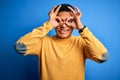 Young handsome latin man wearing yellow casual sweater over isolated blue background doing ok gesture like binoculars sticking Royalty Free Stock Photo