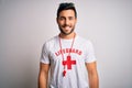 Young handsome lifeguard man with beard wearing t-shirt with red cross and whistle with a happy and cool smile on face