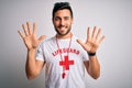 Young handsome lifeguard man with beard wearing t-shirt with red cross and whistle showing and pointing up with fingers number ten