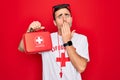 Young handsome lifeguard man wearing t-shirt with red cross and whistle holding first aid kit cover mouth with hand shocked with